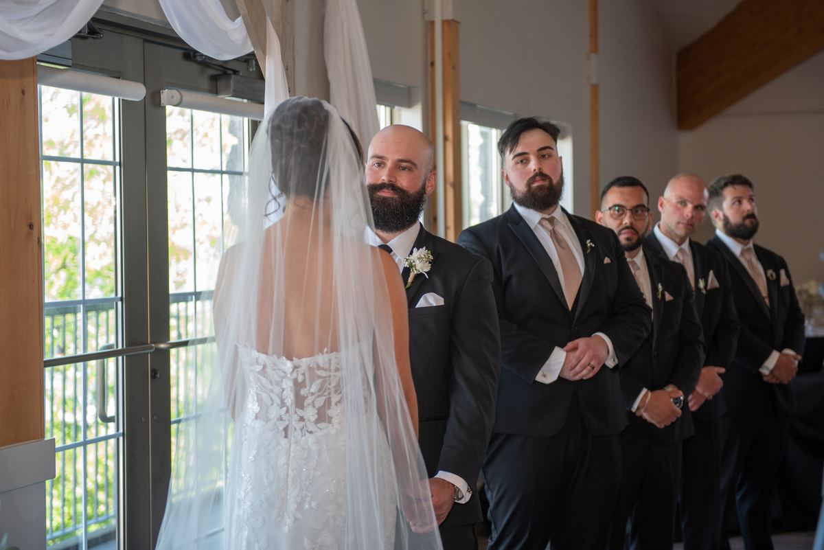 A bride in a lace gown walks down the aisle toward her groom with hands joined, illuminated by natural light.