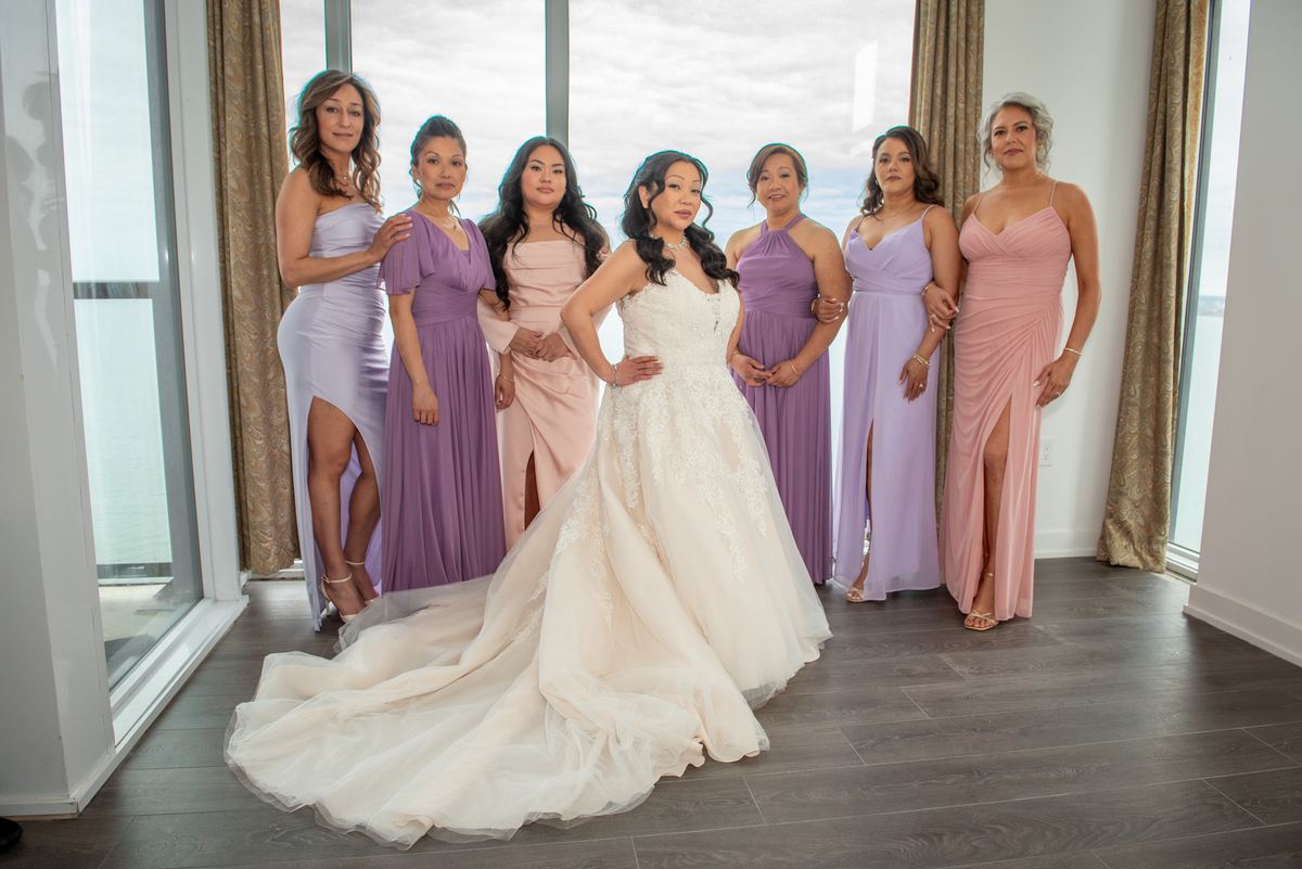 A bride in a white gown stands with her bridesmaids in pastel shades near large windows, bathed in soft daylight.