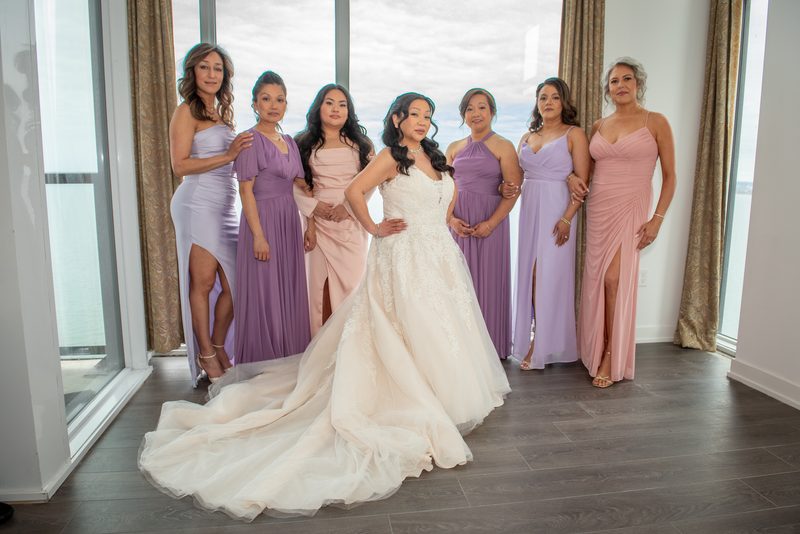 A bride in a white gown stands with her bridesmaids in pastel shades near large windows, bathed in soft daylight.