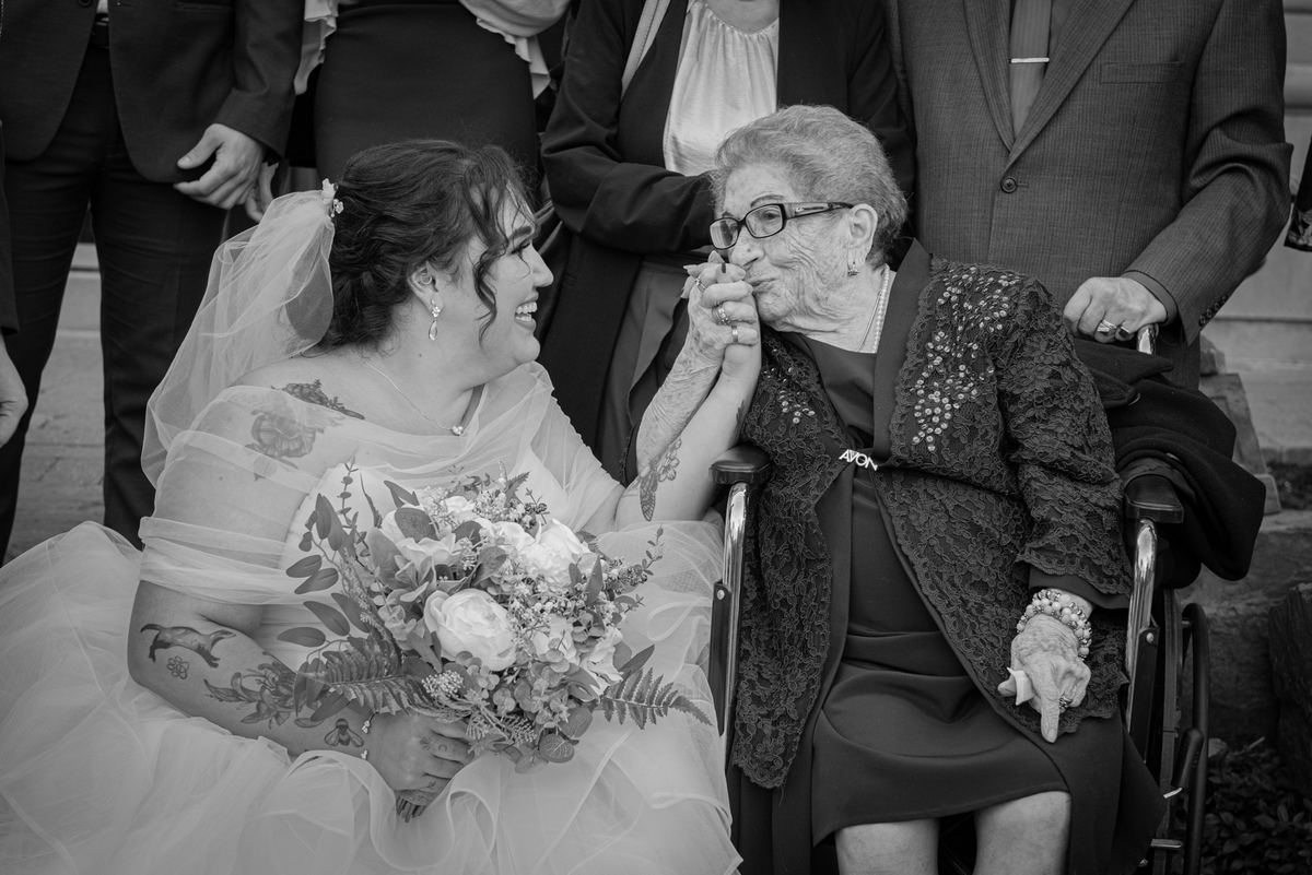 A bride leans toward an elderly woman in a wheelchair, touching hands warmly during a daytime celebration.