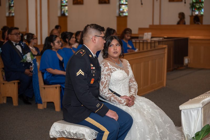 Bride in lace gown and groom in military uniform sit in church pew as light filters through stained glass.
