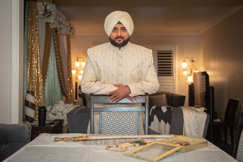 A groom in traditional attire stands behind a chair with ceremonial items on a table in an elegantly decorated room.
