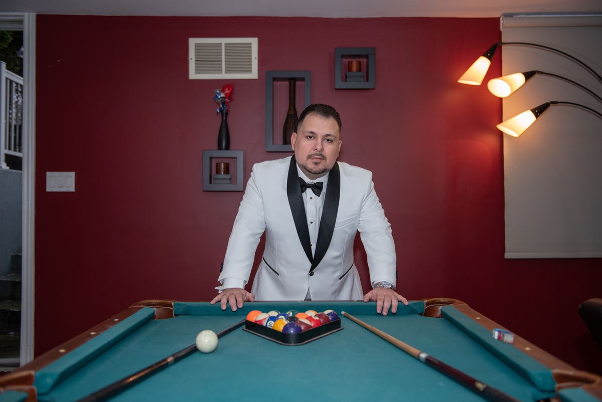 A man in a white tuxedo stands behind a pool table with balls racked, illuminated by a warm floor lamp.