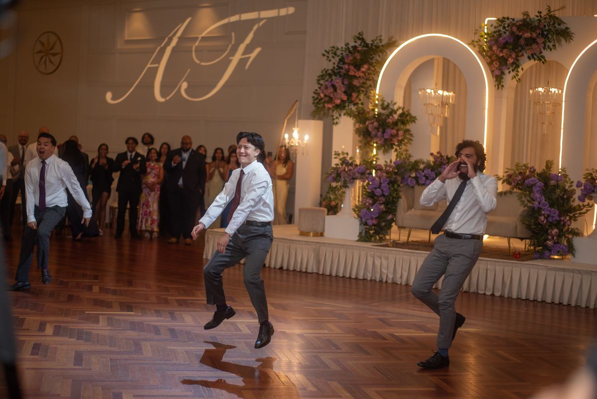 Three men in suits joyfully jump and dance on a wooden floor at a wedding reception, with guests cheering.
