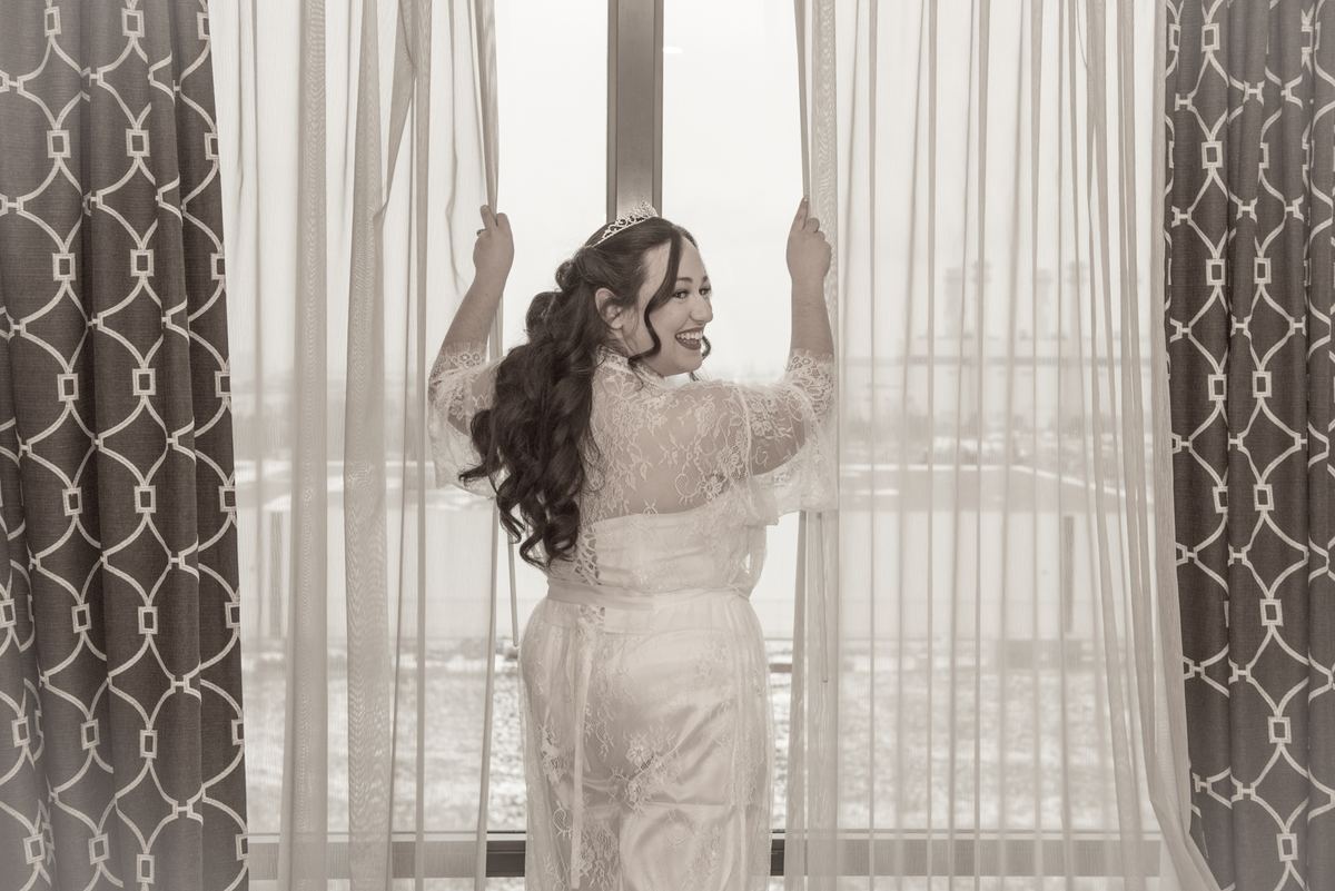 A bride pulls back curtains to reveal a bright view beyond a large window, her expression full of joy.