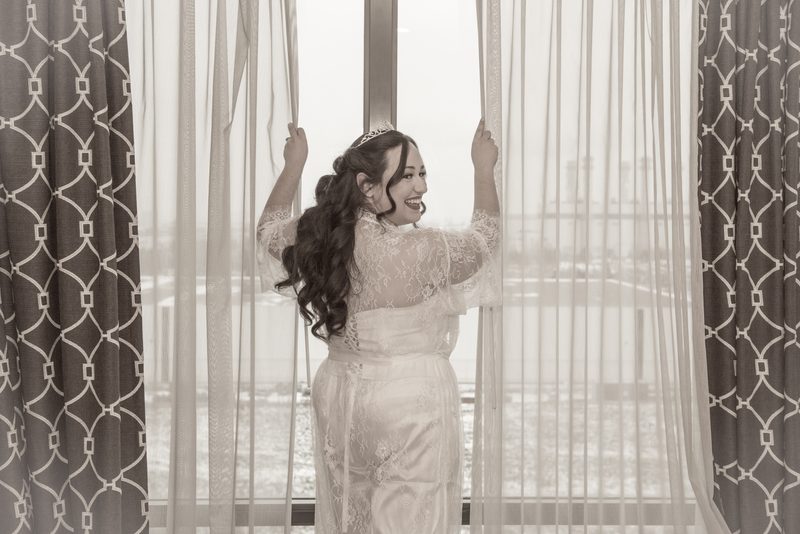 A bride pulls back curtains to reveal a bright view beyond a large window, her expression full of joy.