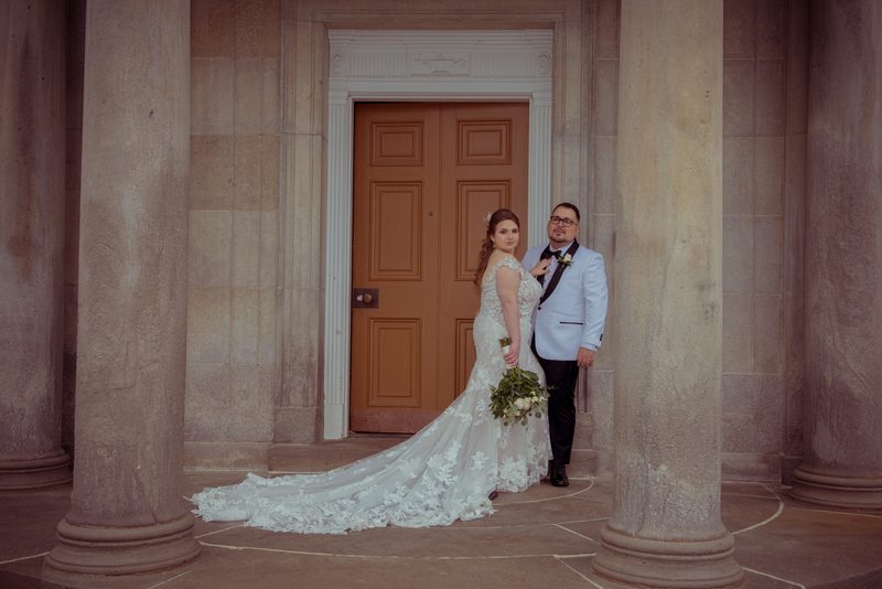 A bride in a lace gown and long train stands with her groom beside an ornate wooden door, hand on shoulder.