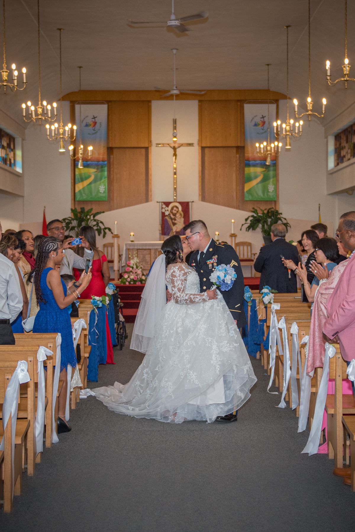 Bride and groom kiss while walking down the aisle surrounded by guests taking photos, under hanging chandeliers.