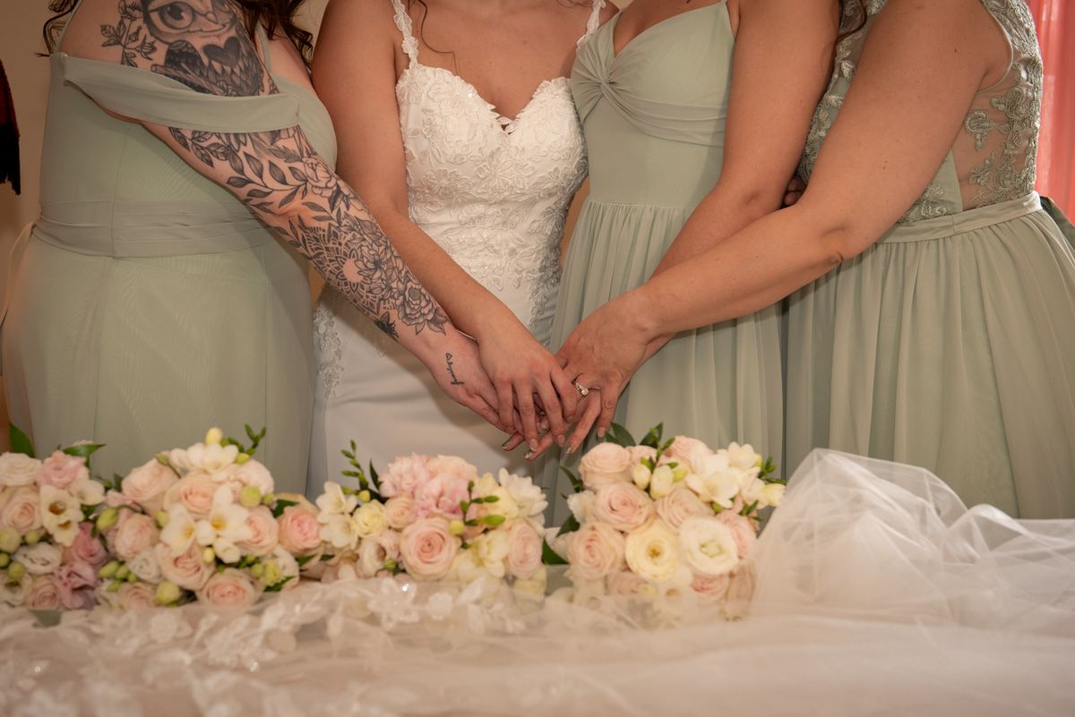 A bride and her bridesmaids stand together, hands clasped, in front of a lace-covered table with floral arrangements.