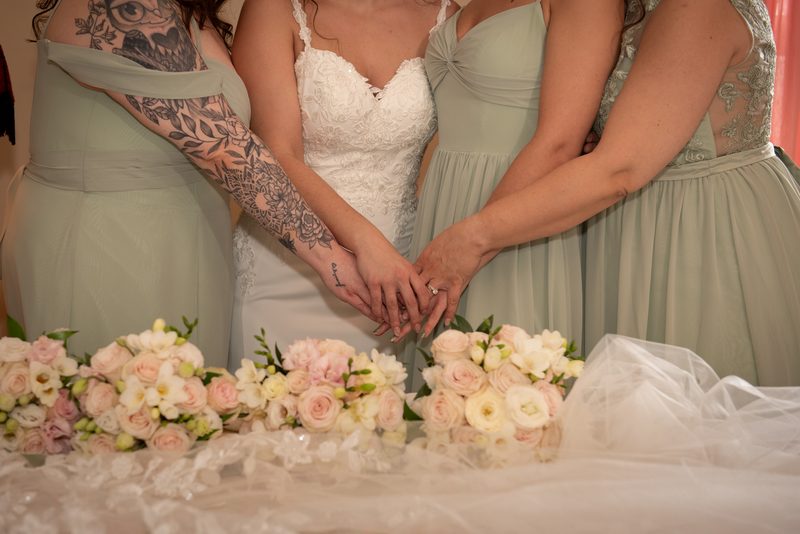 A bride and her bridesmaids stand together, hands clasped, in front of a lace-covered table with floral arrangements.