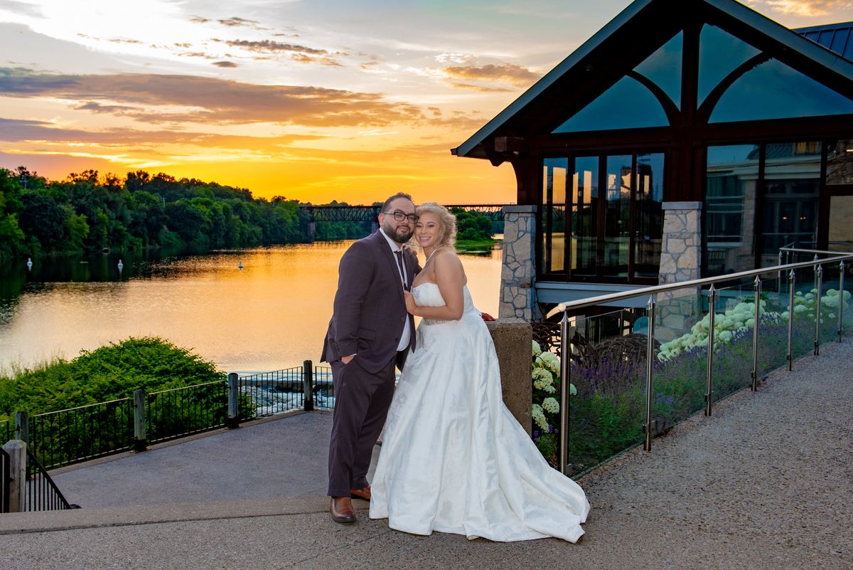 A couple in wedding attire stand close on a riverside patio at sunset, framed by modern architecture and greenery.