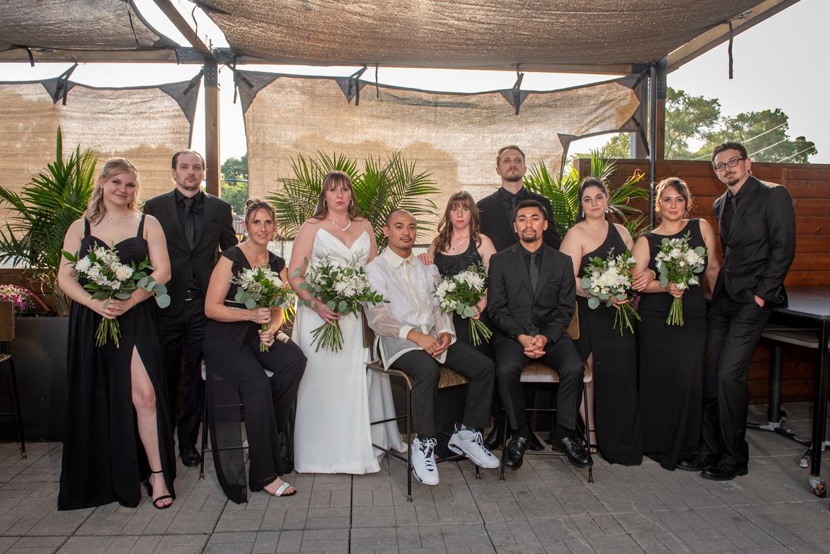 A wedding party poses outdoors under a canopy, the bride and groom in center with bridesmaids in black dresses.