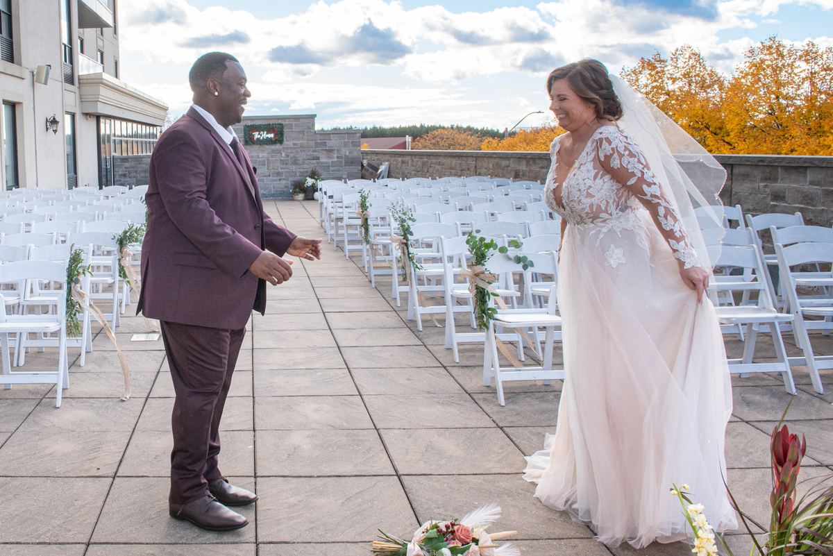 A smiling bride and groom on a stone patio, reaching toward each other as autumn trees frame the background.