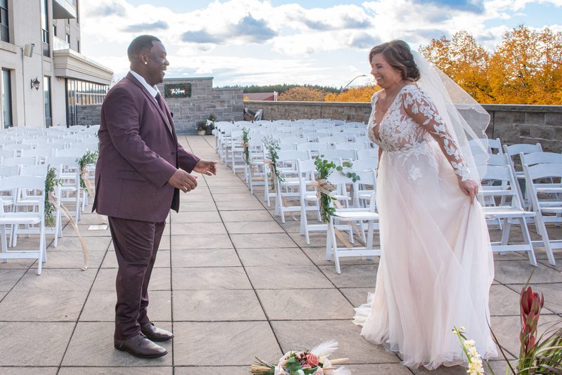 A smiling bride and groom on a stone patio, reaching toward each other as autumn trees frame the background.