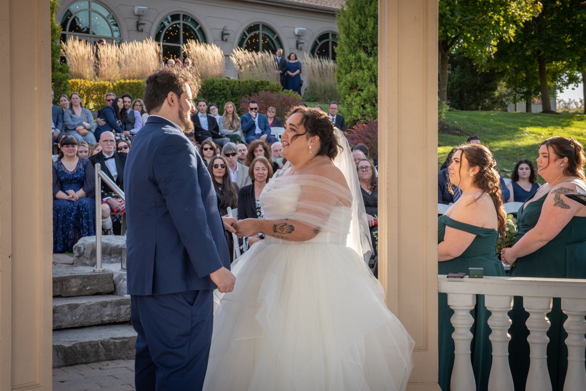 A bride and groom exchange vows under a wooden arch, surrounded by guests and bridesmaids in dark green dresses.