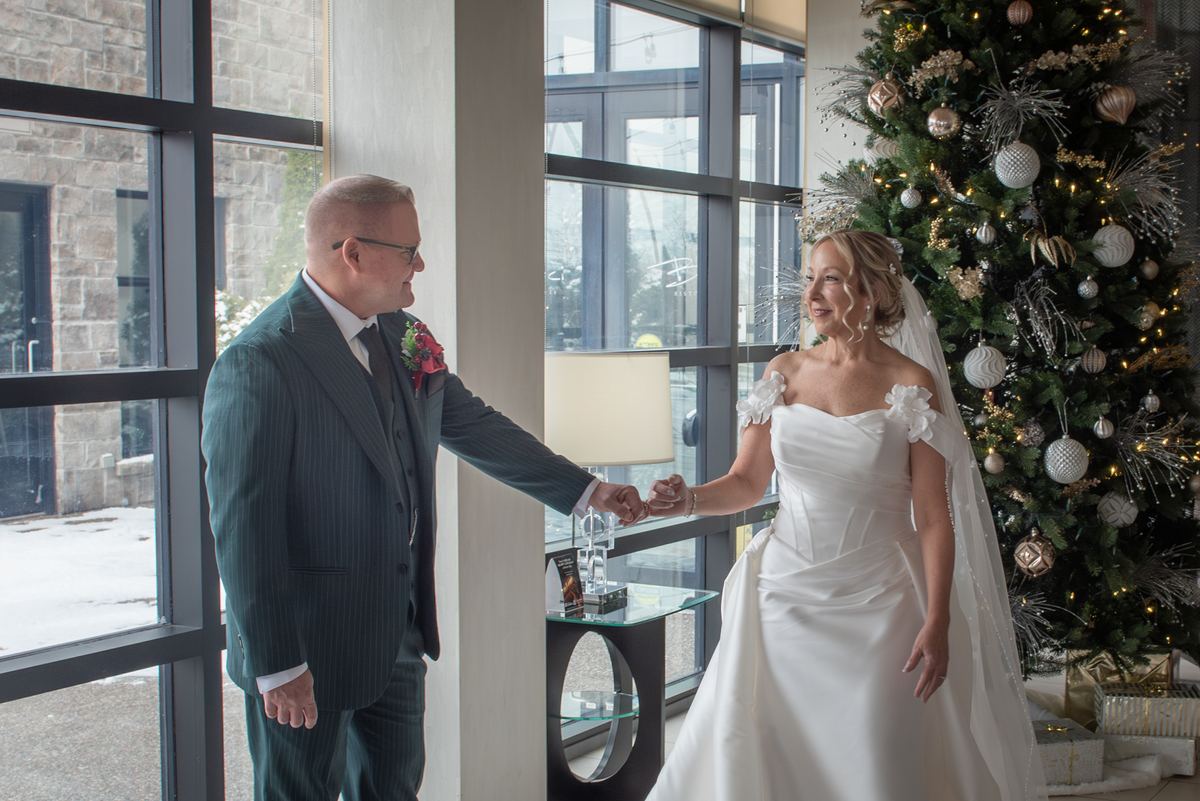 Bride and groom smile and hold hands near a window with a decorated Christmas tree, bathed in natural light.