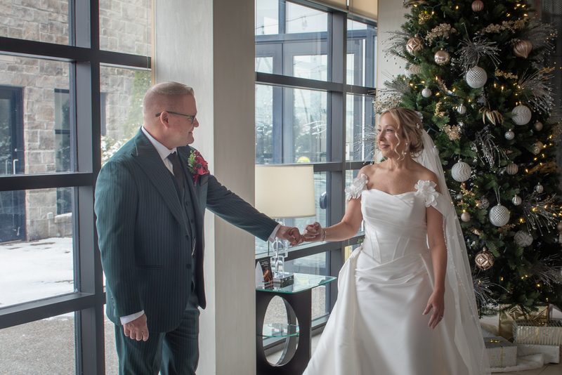 Bride and groom smile and hold hands near a window with a decorated Christmas tree, bathed in natural light.