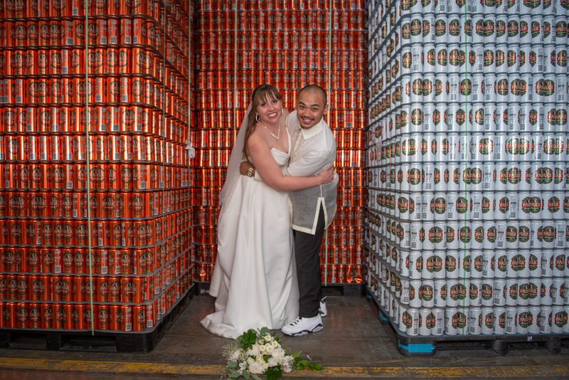 A newlywed couple stands joyfully in a warehouse aisle surrounded by stacks of red and white beer cans, smiling.