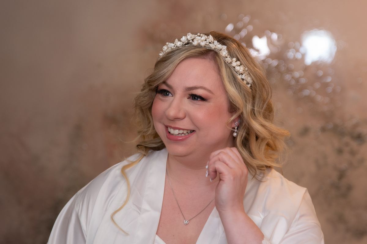 A smiling bride in a white robe and pearl headpiece touches her hair, with soft light and blurred background.
