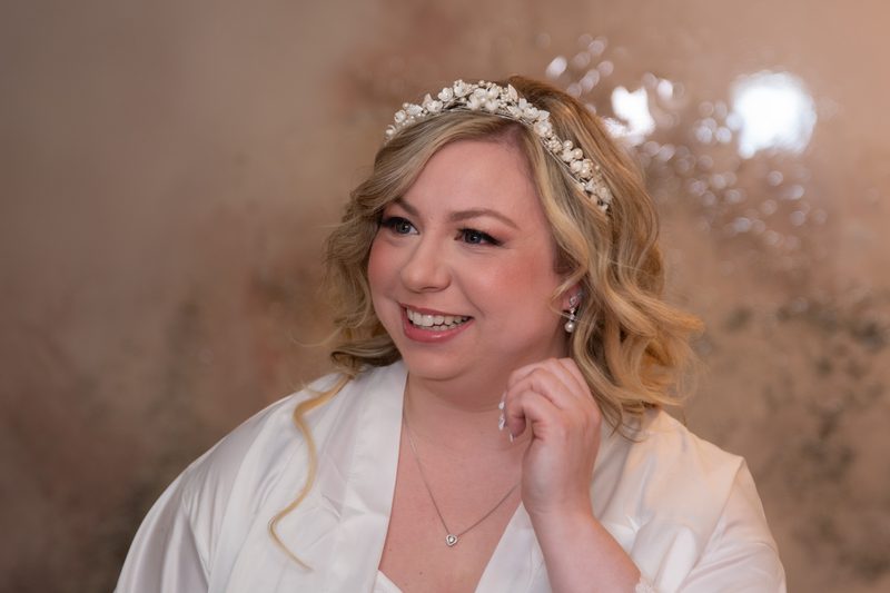 A smiling bride in a white robe and pearl headpiece touches her hair, with soft light and blurred background.