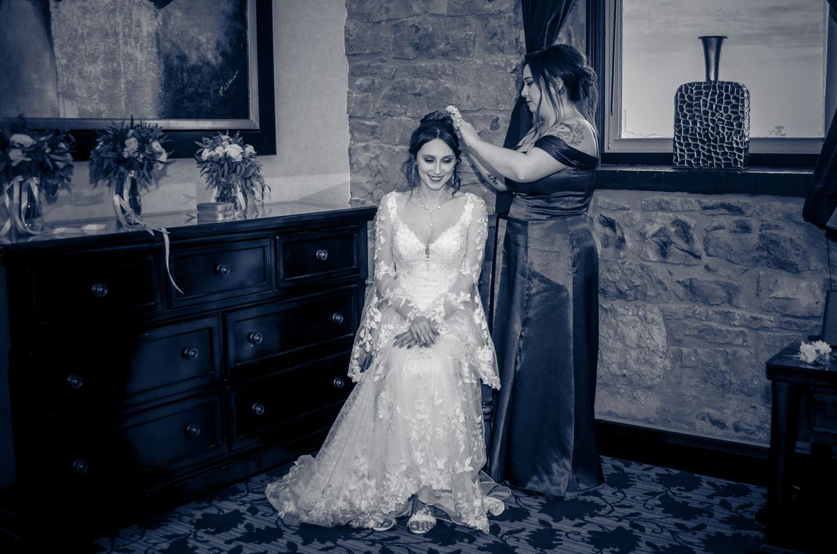 A bride in an ornate lace gown smiles as her attendant adjusts her hair in a softly lit room with stone walls.