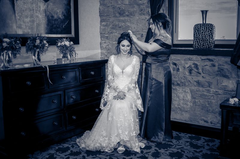 A bride in an ornate lace gown smiles as her attendant adjusts her hair in a softly lit room with stone walls.