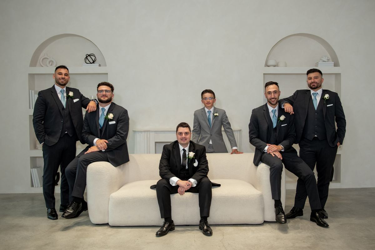 A group of six men, including groom Vince, pose in suits with boutonnieres near a white sofa in a modern room.