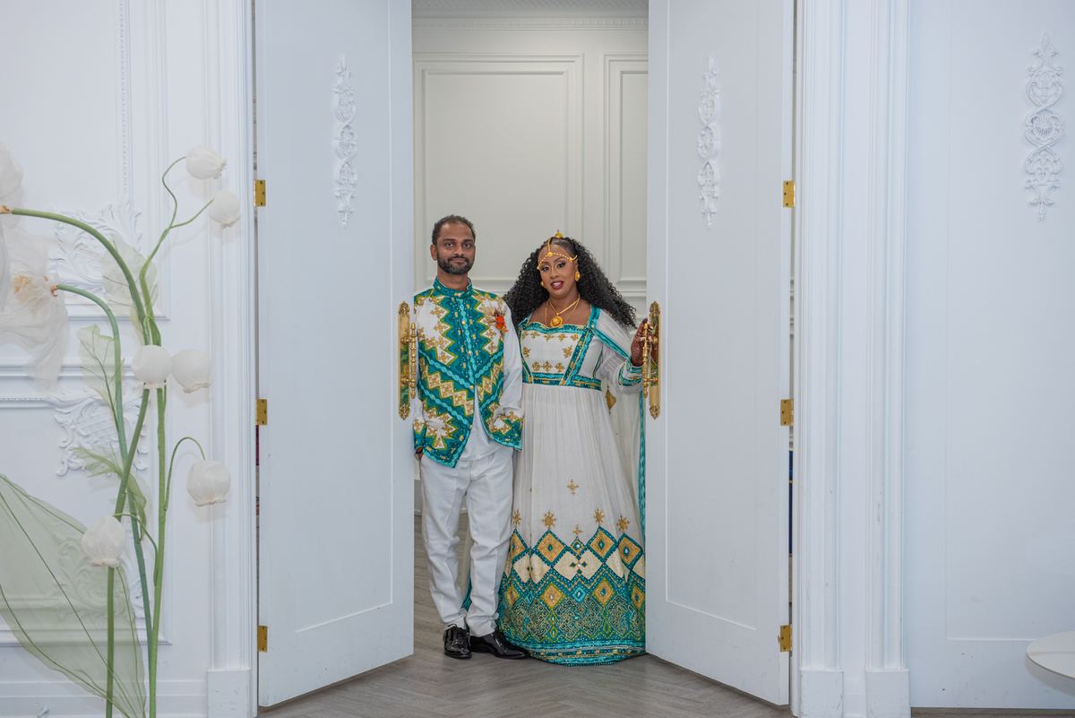 A couple in ornate traditional attire stands in an elegant doorway with gold embroidery and floral decor.