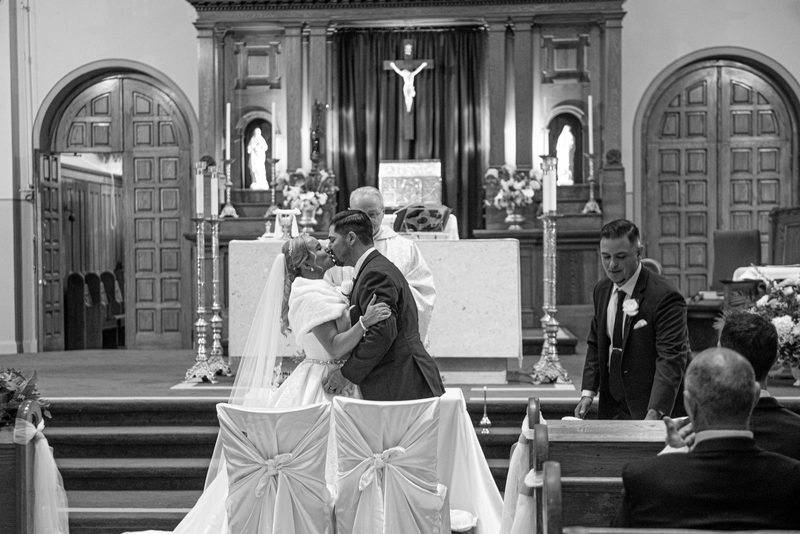 A bride and groom kiss at an altar surrounded by attendees, with arched doorways and religious iconography in the background.