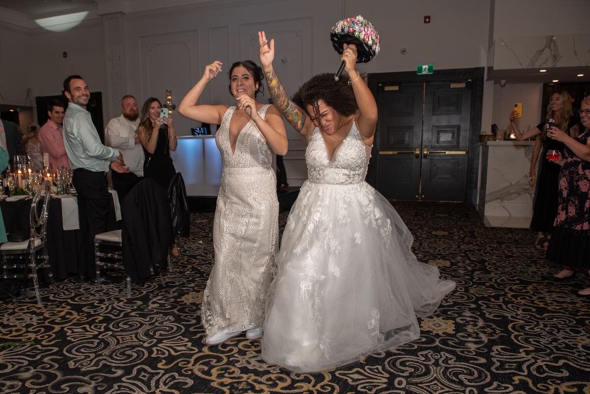 Two brides celebrate at a reception, one holding her bouquet aloft while the other dances beside her.