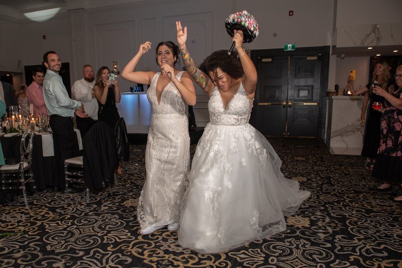 Two brides celebrate at a reception, one holding her bouquet aloft while the other dances beside her.