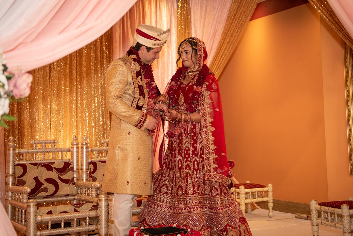 A bride and groom in traditional Indian attire exchange rings under ornate chairs and golden-red drapery.
