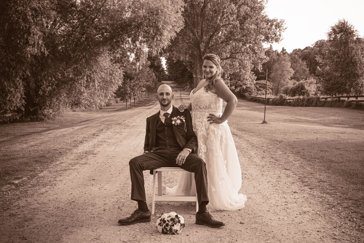 A newlywed couple poses by a tree-lined dirt road, the bride smiling and touching her groom's shoulder.