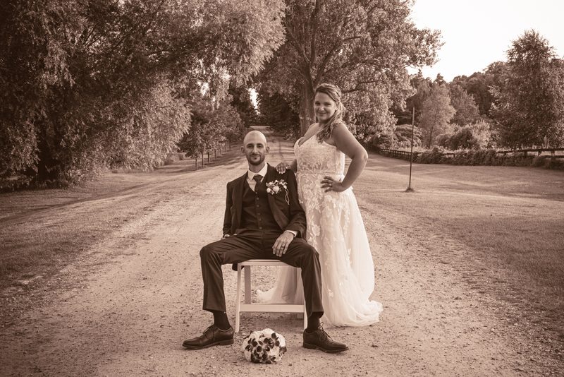 A newlywed couple poses by a tree-lined dirt road, the bride smiling and touching her groom's shoulder.