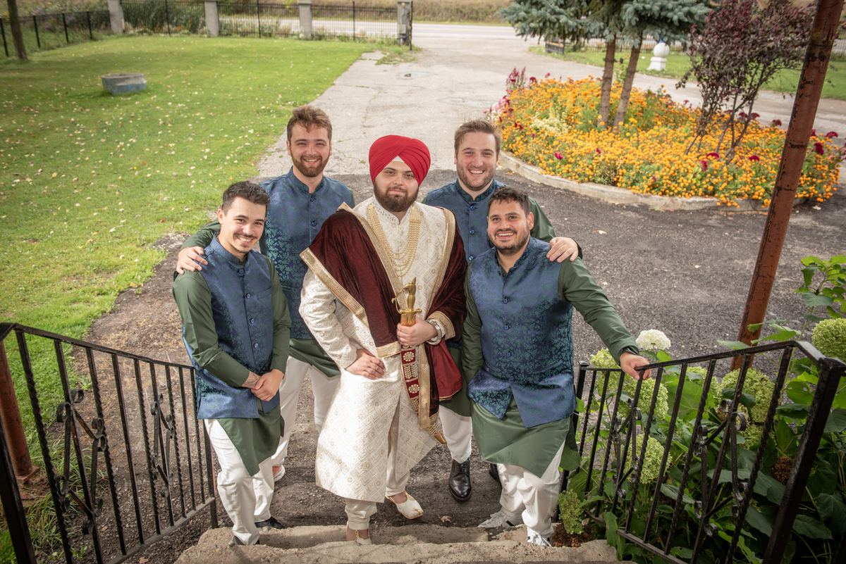 A groom in a white sherwani and red turban with four smiling men on outdoor stone steps, holding a ceremonial kirpan.