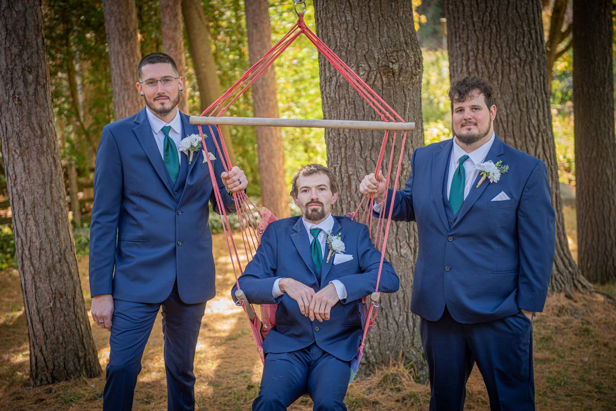 Three men in navy suits pose with one sitting in a red macrame swing held by the others in a sunlit forest, smiling.