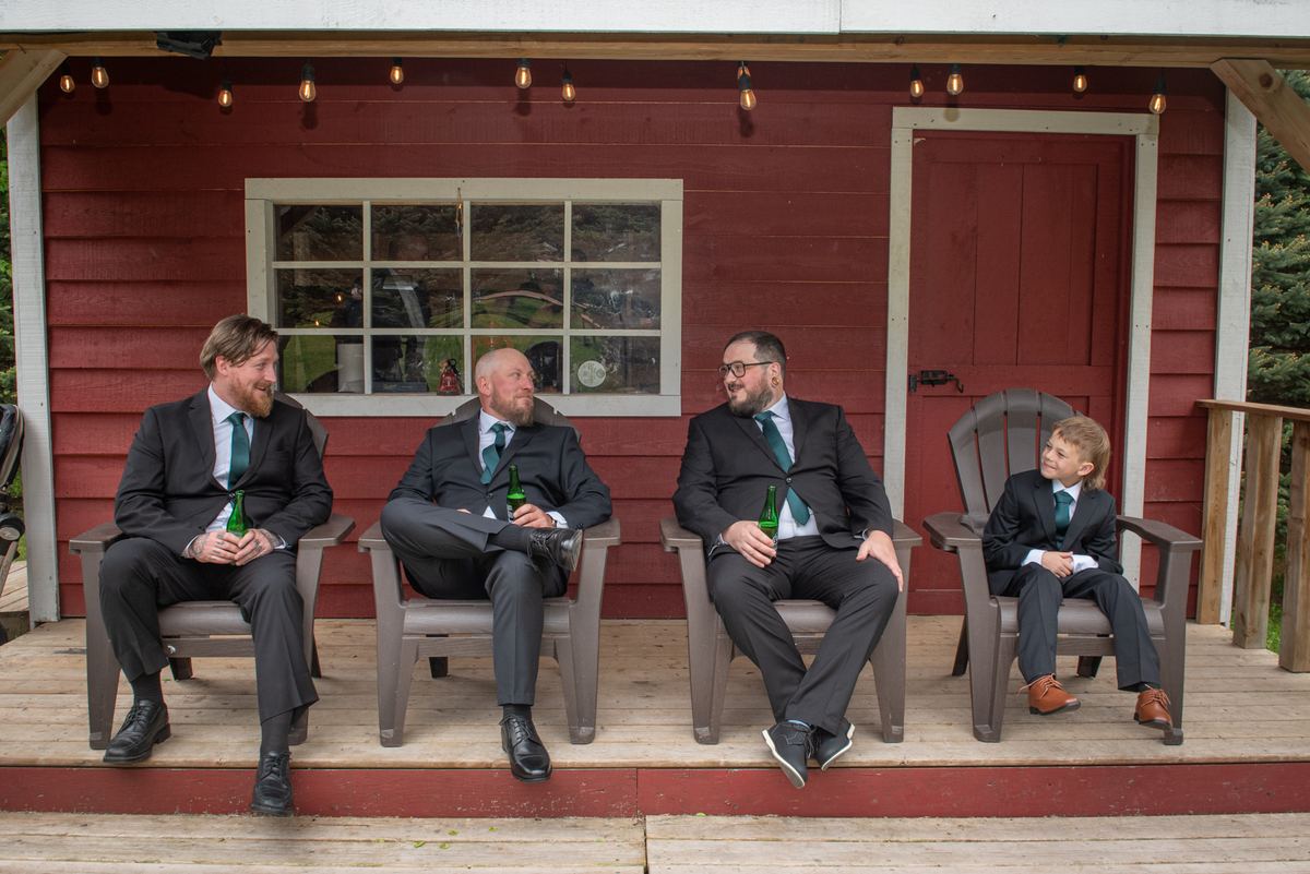 Four men and a young boy in dark suits and green ties sit on a wooden porch with beer bottles, illuminated by natural light.