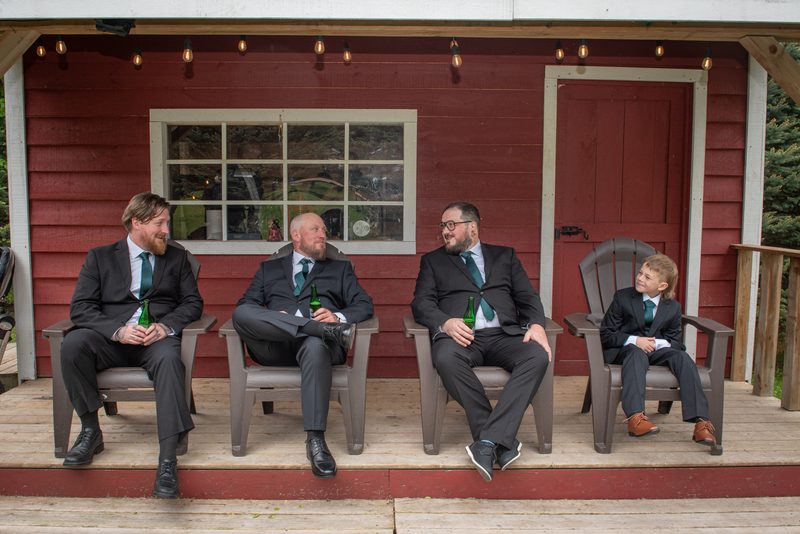 Four men and a young boy in dark suits and green ties sit on a wooden porch with beer bottles, illuminated by natural light.