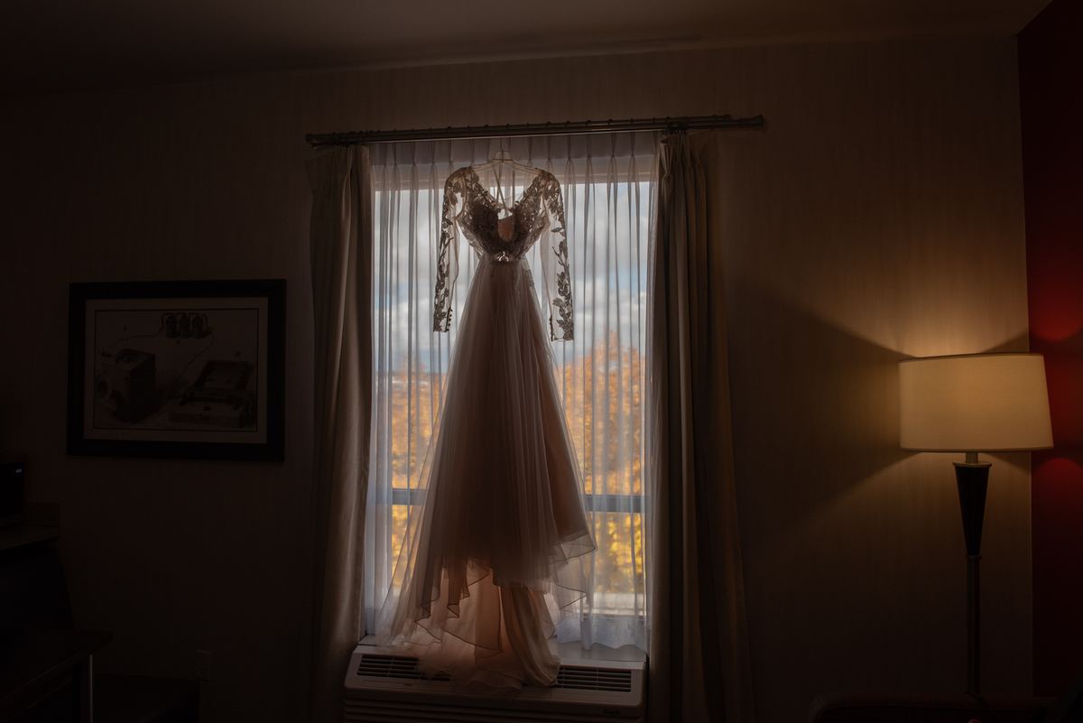A wedding gown hangs in front of a window, illuminated by golden late afternoon light with autumn trees visible outside.