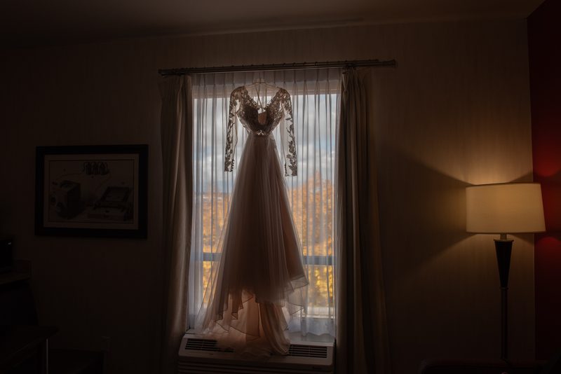 A wedding gown hangs in front of a window, illuminated by golden late afternoon light with autumn trees visible outside.