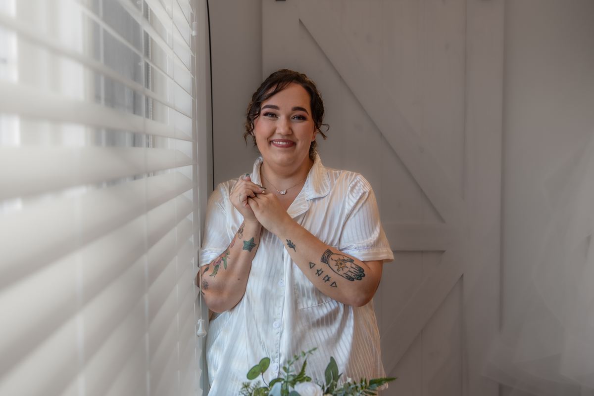 A woman with tattoos smiles by a window, hands clasped, in a white shirt and necklace, with greenery nearby.