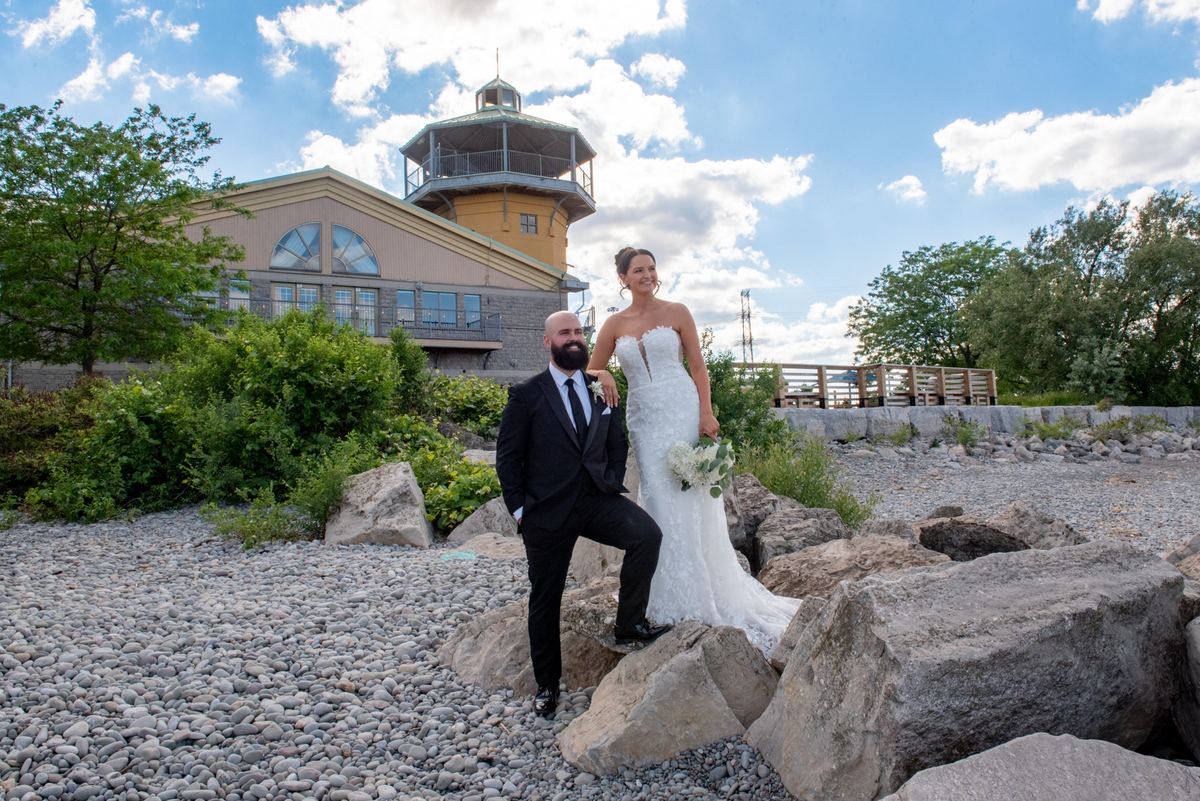 Kara and Tiago pose together on a rocky shoreline, with the distinctive yellow lighthouse-style building in the background.