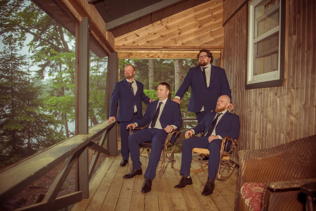 Four men in navy suits stand casually on a wooden porch overlooking a misty forest, bathed in soft golden light.