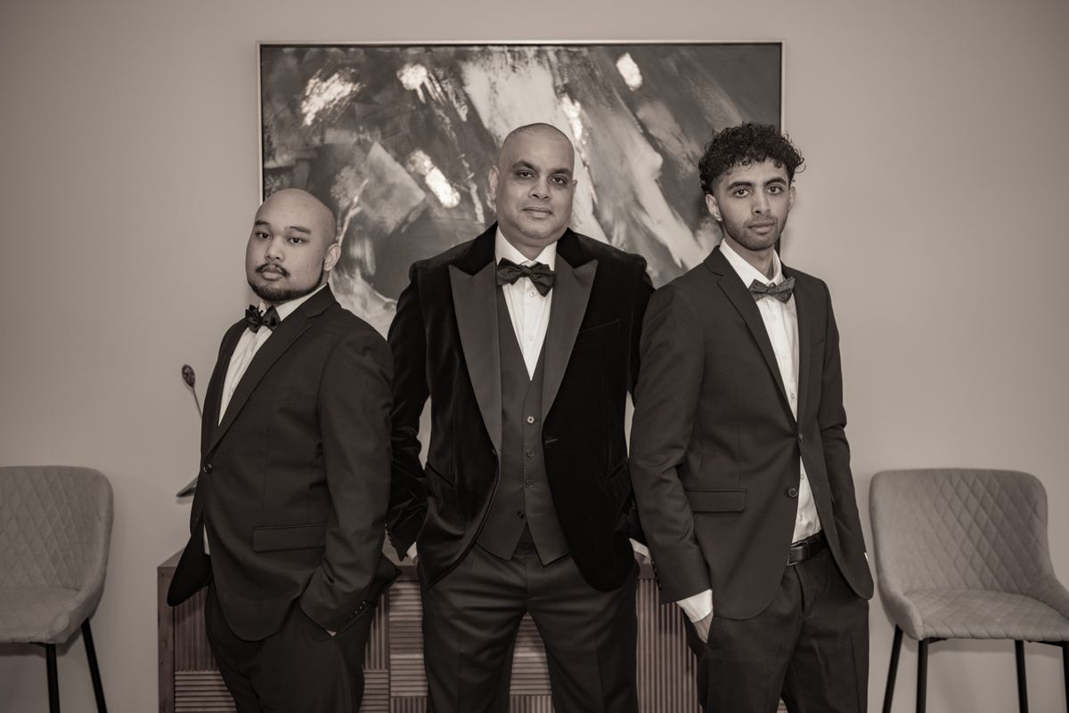 Three men in tuxedos stand confidently for a portrait against an abstract painting and chairs, in warm sepia tones.