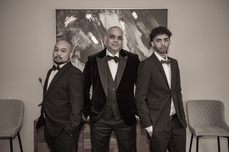 Three men in tuxedos stand confidently for a portrait against an abstract painting and chairs, in warm sepia tones.