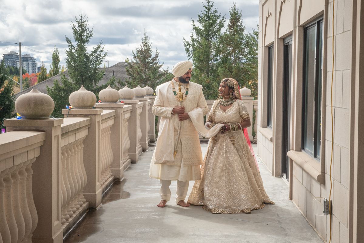 Bride and groom in traditional South Asian attire walk hand-in-hand on stone balcony under overcast sky.