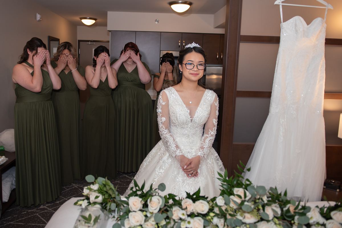 A bride in a white lace gown stands with her back to the camera, surrounded by bridesmaids in olive green dresses.
