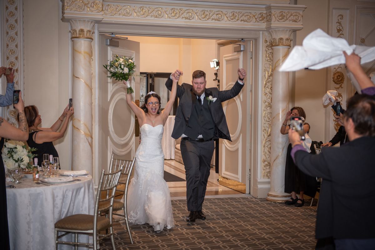 A bride and groom joyfully enter a decorated banquet hall, arms raised as guests cheer and capture the moment with phones.