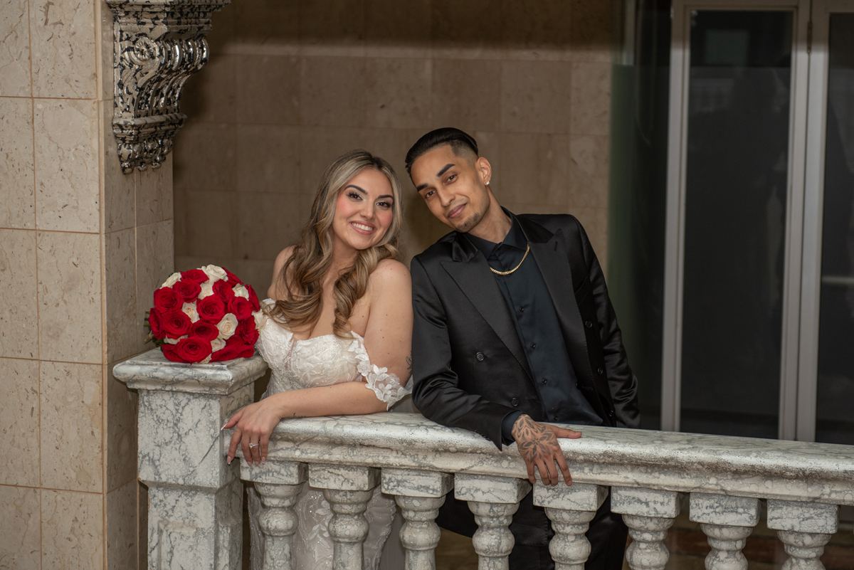 A smiling bride and groom pose together on a marble balcony, the bride holding a red-and-white rose bouquet.