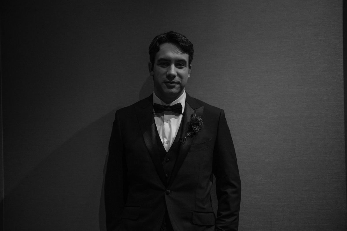 A man in formal wedding attire stands against a dimly lit wall, illuminated by soft light, calm and composed.
